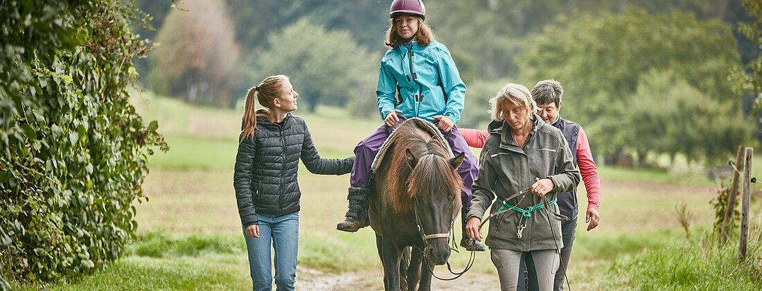Bild: Kind sitzt auf dem schwankenden Rücken eines Pferdes und wird von Eltern und Therapeuten gestützt und begleitet.
